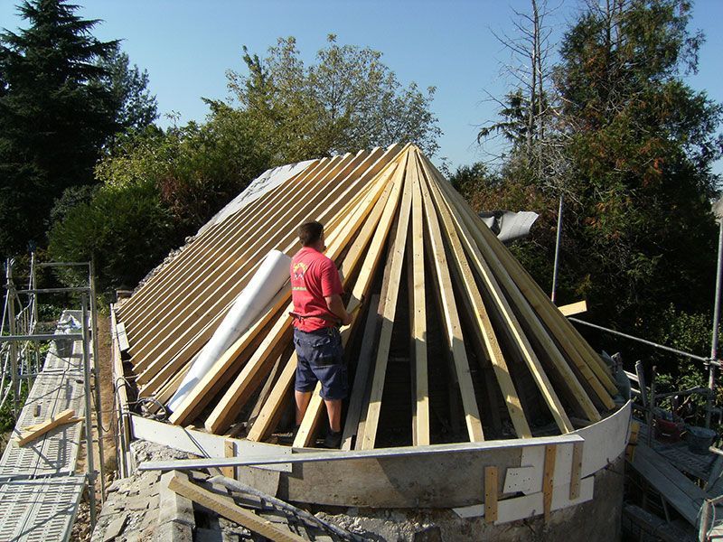 Ouvrier debout sur un bâtiment circulaire en construction, doté d'une charpente de toit conique en bois.