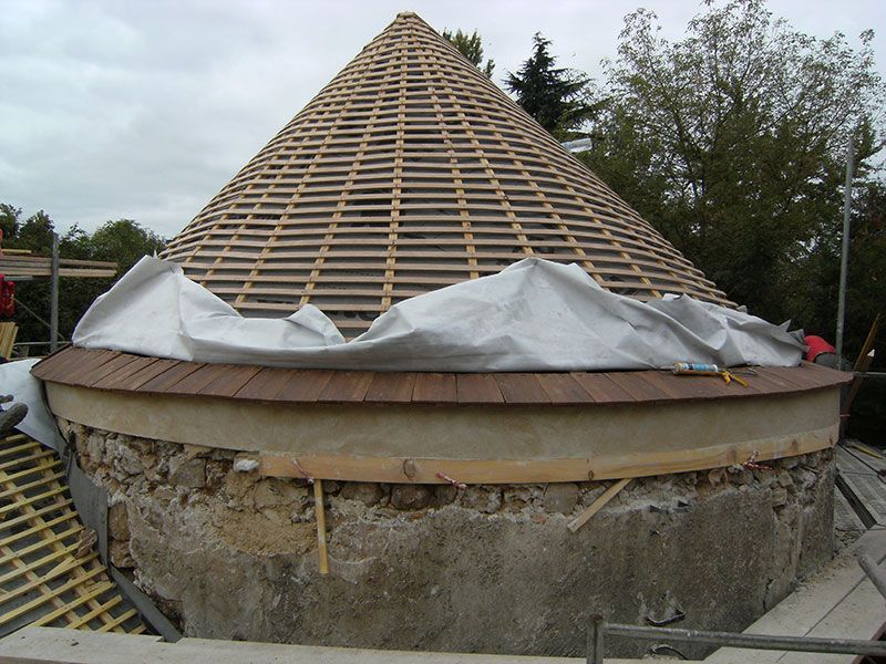 Structure circulaire en pierre en construction avec une charpente de toit conique en bois et une bâche blanche drapée autour.