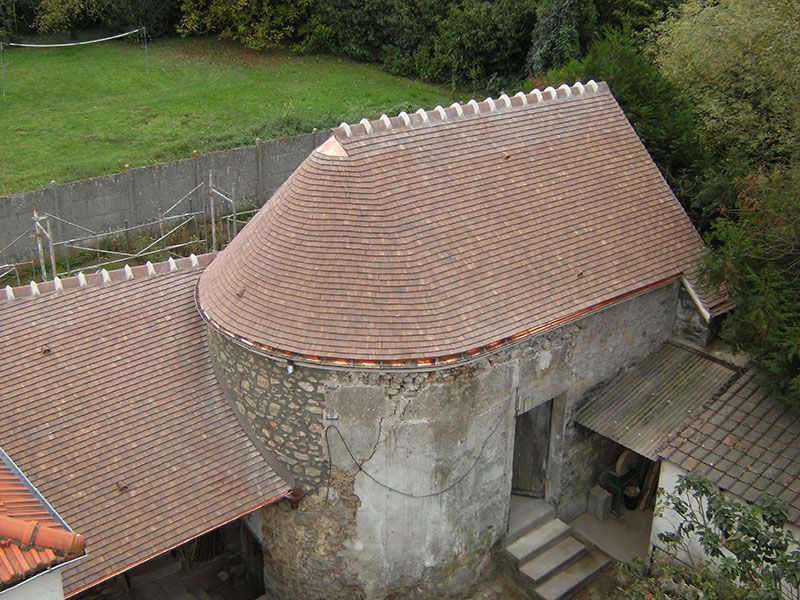 Vue aérienne d'un bâtiment en pierre avec un toit arrondi en tuiles rouges et des marches, entouré de verdure.