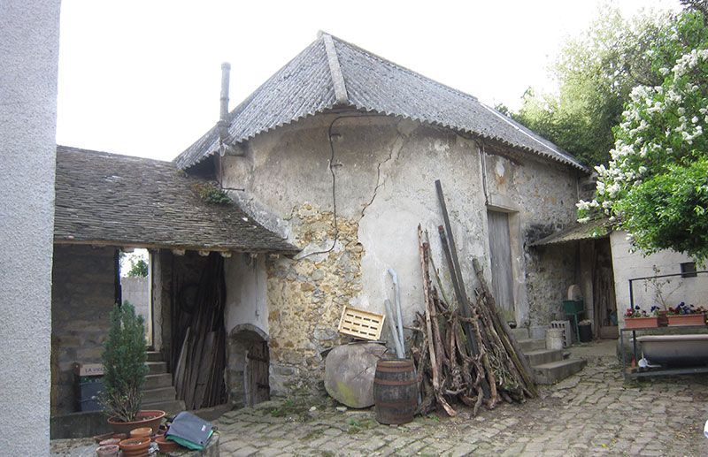 Cour d'une ancienne ferme en pierre avec du bois de chauffage empilé, un tonneau et un sol pavé