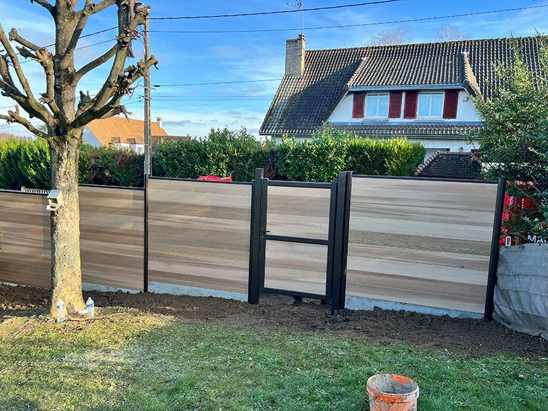 Clôture en bois dans une cour avant avec un arbre, de l'herbe et une maison derrière, sous un ciel bleu.