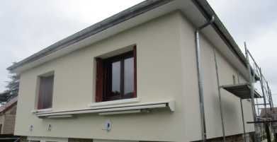 Maison beige à deux étages avec des boiseries foncées et un auvent de fenêtre, vue de l'extérieur avec un échafaudage sur la droite