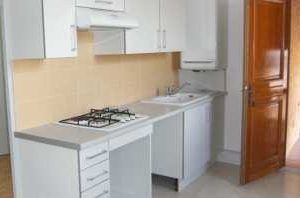 Petite cuisine avec placards blancs, crédence beige, plaque de cuisson, évier et porte en bois ouverte sur la droite.