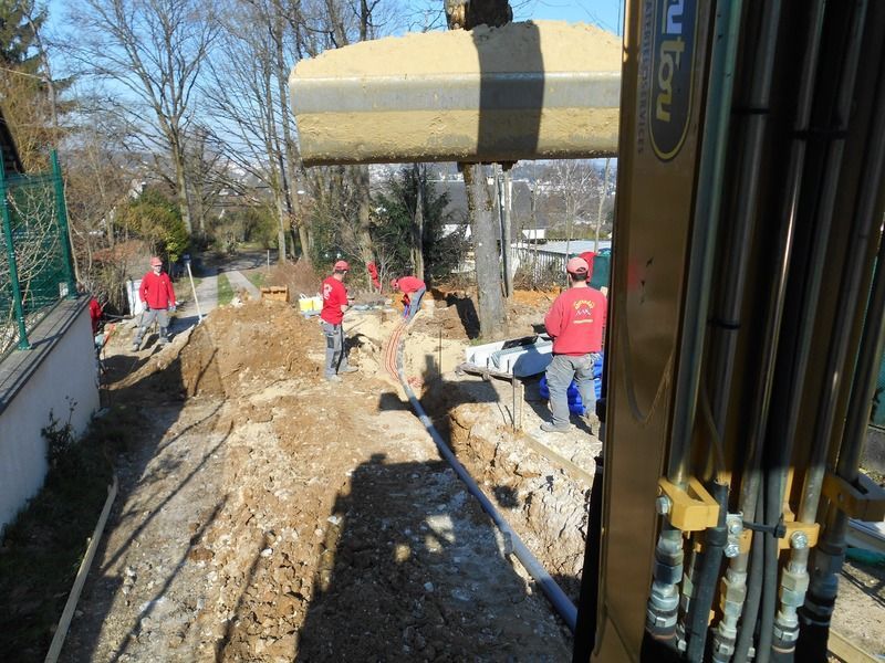 Des ouvriers en chemises rouges creusent une tranchée à côté d'une grande excavatrice sur un chantier boueux.