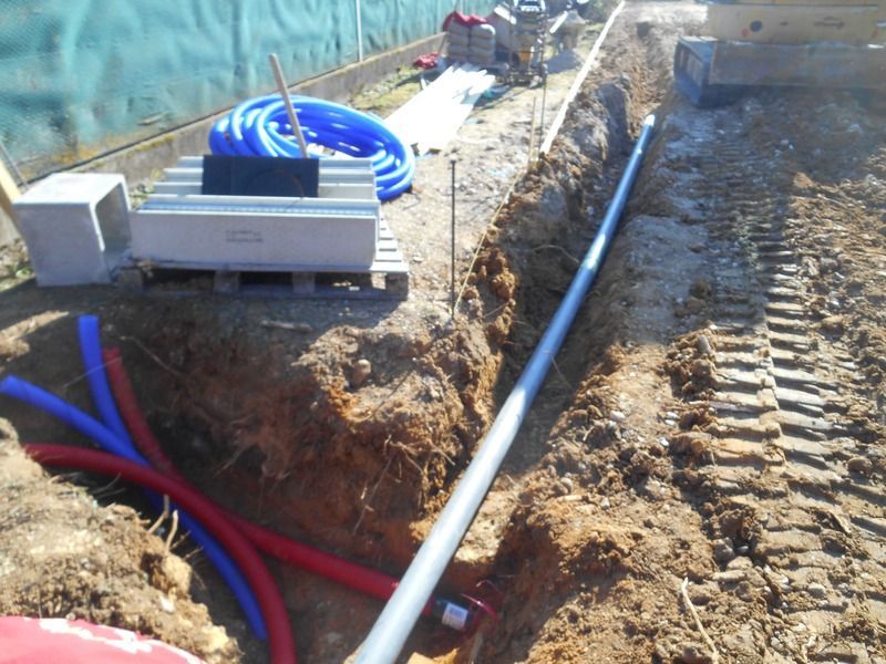 Tranchée de chantier avec canalisations enterrées, tuyaux bleus et rouges, et chenille d'excavatrice au bord d'un chemin de terre.