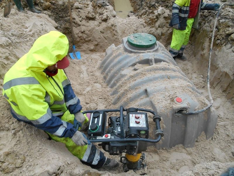 Des ouvriers vêtus de cirés fluo utilisent des engins autour d'une cuve souterraine enterrée sur un chantier de construction.