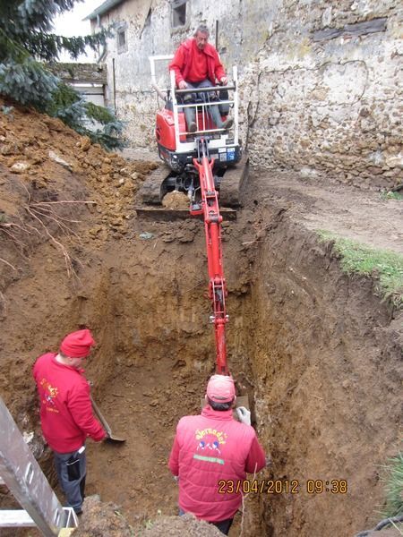 Un ouvrier manœuvrant le bras d'une excavatrice rouge dans une tranchée profonde à côté d'un mur en béton