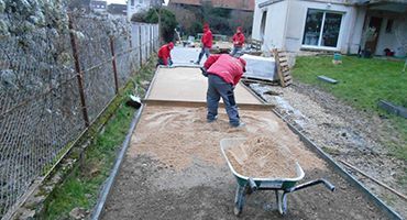 Des ouvriers nivellent le sol dans une cour arrière, avec une brouette au premier plan et une terrasse à côté d'une maison.