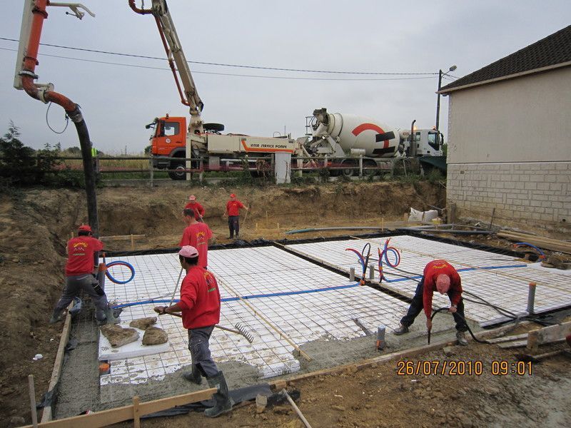 Des ouvriers en vestes rouges coulent du béton sur un chantier de fondation à l'aide d'une pompe à béton placée au-dessus.