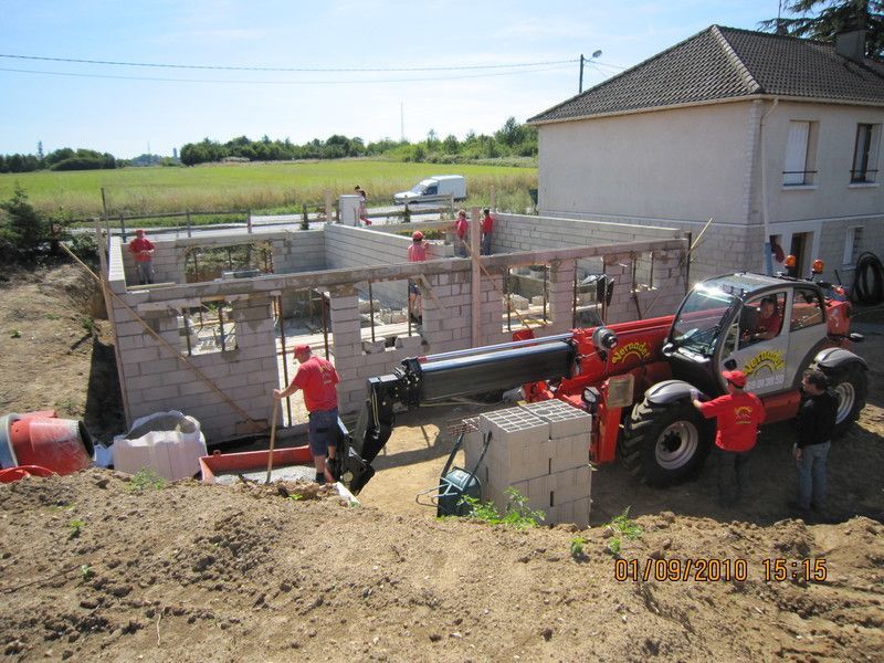 Des ouvriers et un chariot télescopique rouge sur un chantier de construction à côté d'une maison, en train de bâtir des murs en pierre.