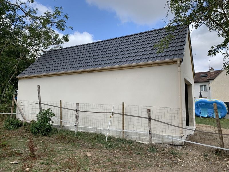 Petite maison blanche au toit gris foncé, derrière une clôture en fil de fer.