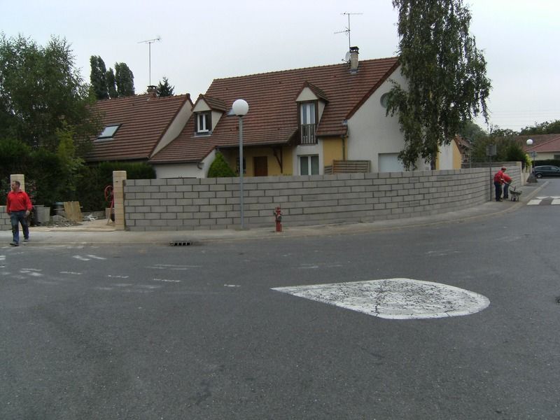 Coin de rue résidentielle avec des maisons derrière un muret en pierre et une personne marchant sur la gauche.