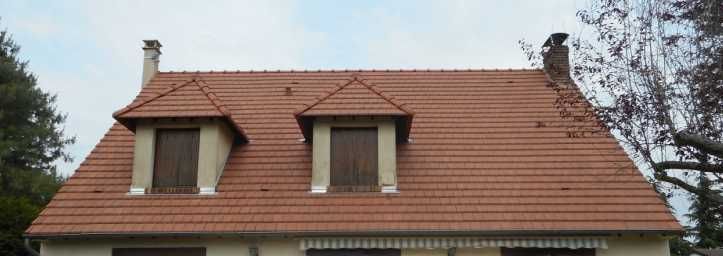 Maison au toit de tuiles rouges avec deux lucarnes et des cheminées sous un ciel nuageux