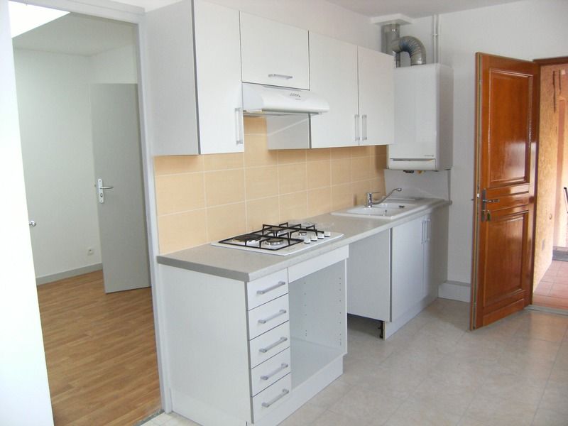 Petite cuisine blanche avec cuisinière à gaz, évier, placards et porte en bois ouverte sur la droite.