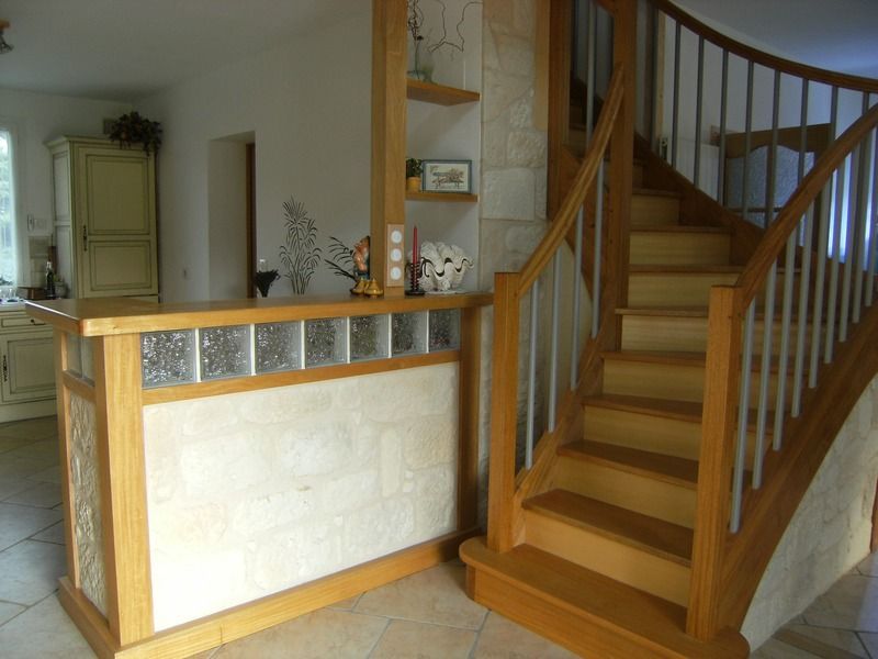 Un escalier en bois jouxte une petite cloison dans l'entrée d'une maison, avec des pavés de verre et une plante en arrière-plan.