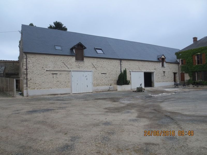 Ferme en pierre au toit gris et aux portes blanches, située dans une cour tranquille sous un ciel couvert.