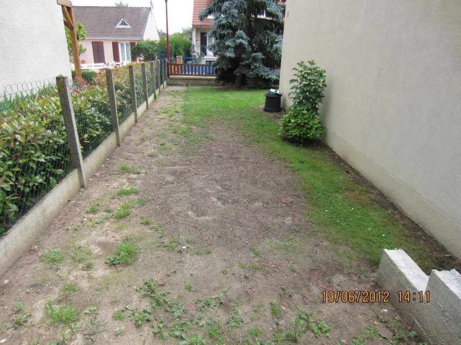 Cour latérale étroite en terre battue entre une maison et un mur, avec une pelouse clairsemée, une clôture et un portail au fond.