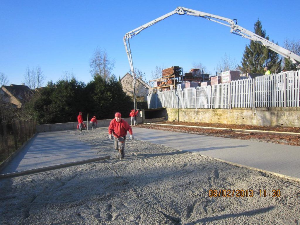 Des ouvriers coulent du béton sur une allée en pente à côté d'une maison, avec un camion-pompe à béton au-dessus.