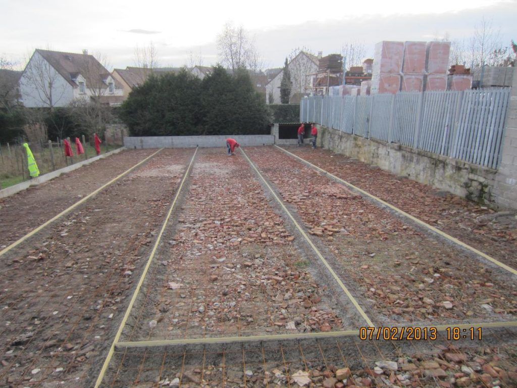 Chantier de construction en plein air avec de longues allées de gravier, des bordures en bois et des ouvriers à proximité de maisons et d'une clôture.