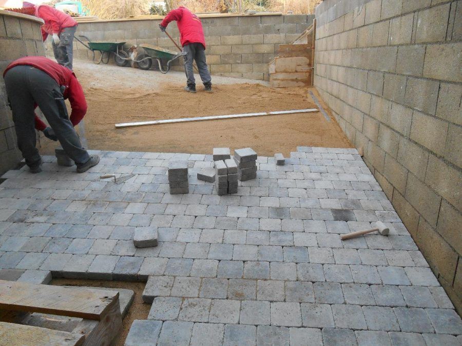 Des ouvriers posent des pavés gris dans une cour entourée de murs de briques, avec des blocs empilés au sol.