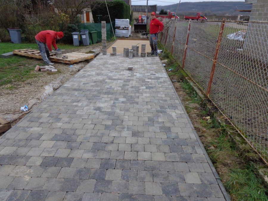 Des ouvriers posent des pavés gris sur un trottoir longeant un chemin clôturé à l'extérieur.