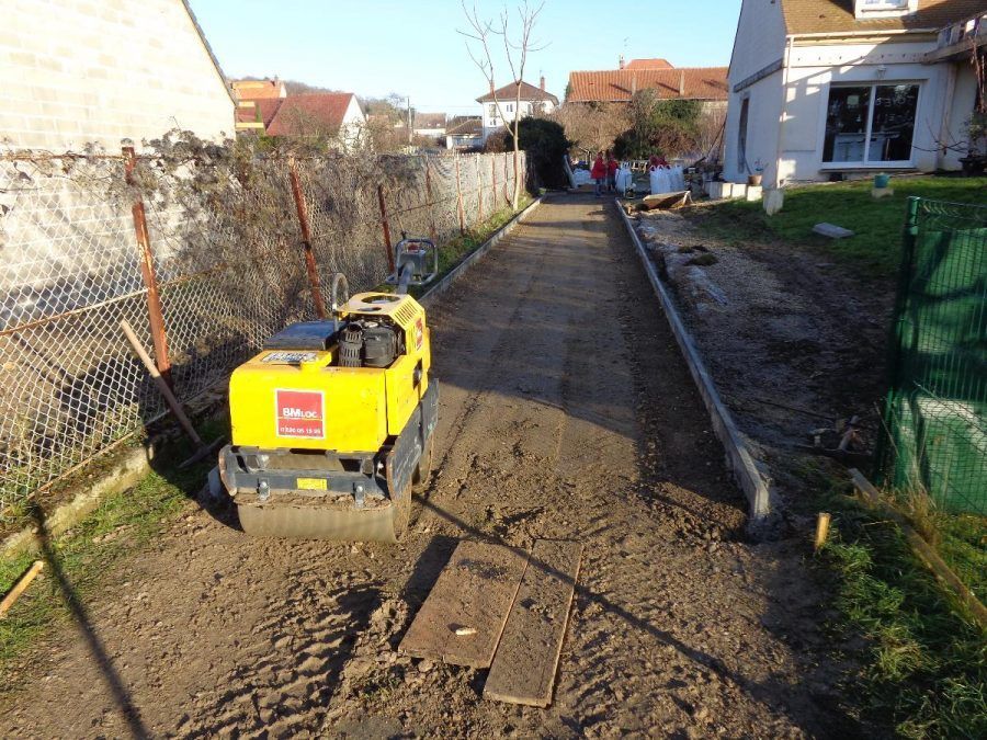 Un compacteur de chantier jaune se trouve sur un chemin de terre entre des maisons, avec une tranchée et une clôture à proximité.