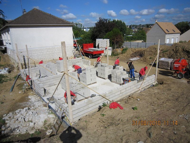 Chantier de construction avec murs en béton en construction, ouvriers, engins rouges et deux maisons en arrière-plan