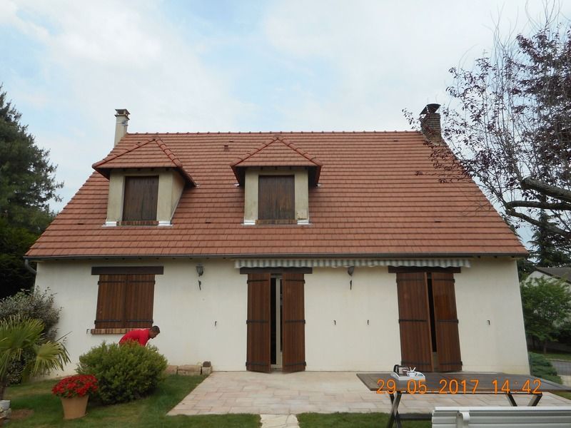 Petite maison beige avec un toit de tuiles rouges, des volets marron et un jardin à l'avant.