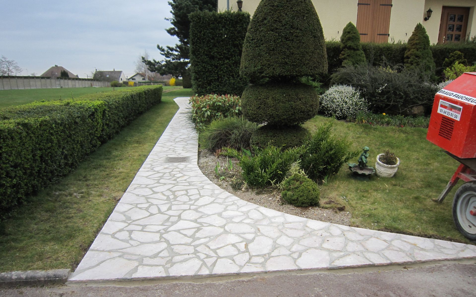 Allée de jardin en pierre bordée de haies et d'arbustes taillés, avec une boîte aux lettres rouge sur la droite.