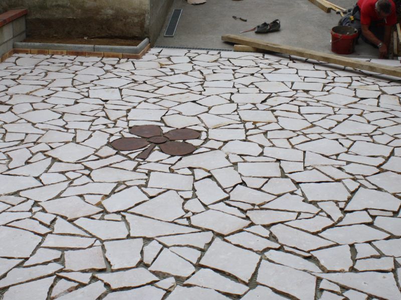 Terrasse en pierre blanche irrégulière avec un petit motif circulaire brun au centre, bordée de béton et d'outils.