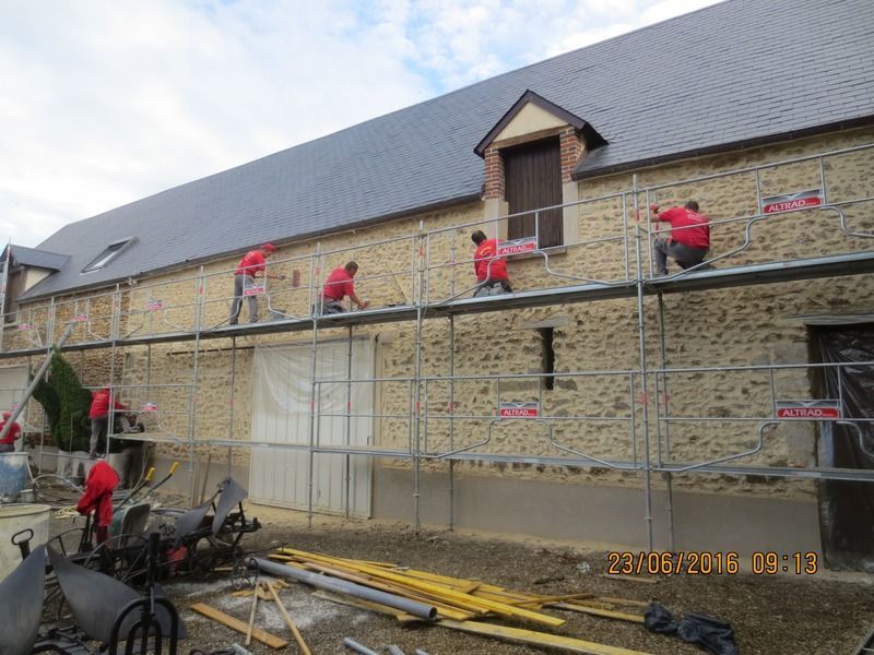Des ouvriers en vestes rouges réparent, sur des échafaudages, le mur d'un bâtiment en pierre sous un toit gris.