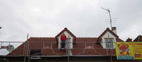 Toit en tuiles rouges avec deux lucarnes, une antenne de télévision et une enseigne colorée sur la droite.