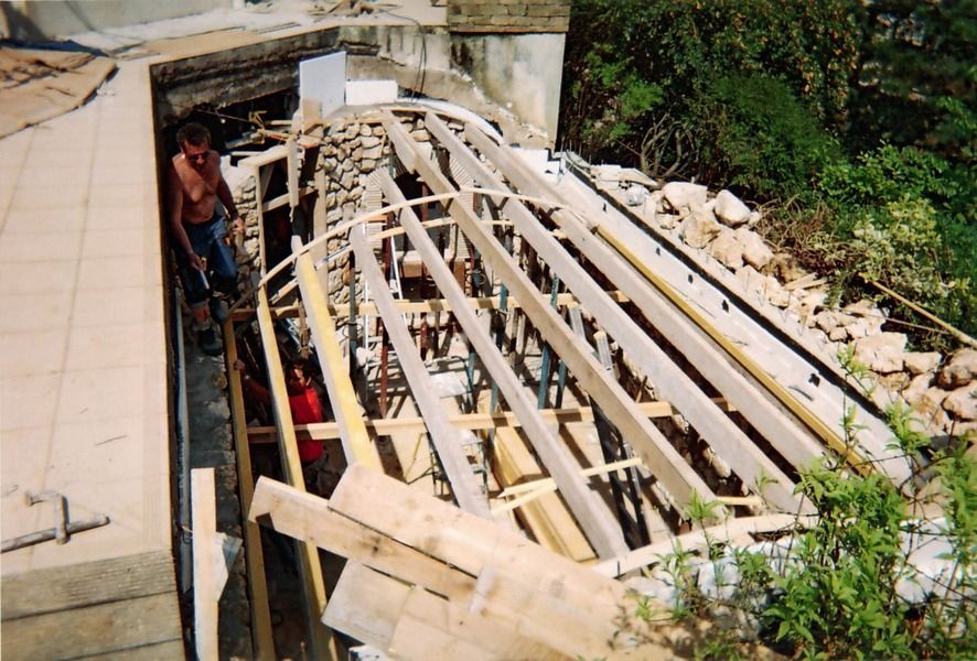 Un ouvrier se tient debout à côté de fermes de toit en bois en construction, près d'un mur de pierre et de verdure.