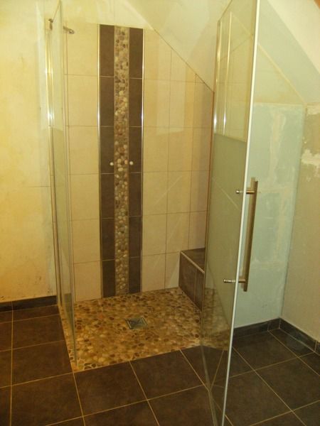 Douche à l'italienne avec portes en verre, murs carrelés beiges, carrelage foncé au sol et receveur de douche en galets.