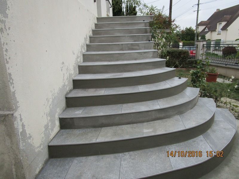 Escalier extérieur incurvé en béton, adossé à un mur blanc et montant vers la droite.