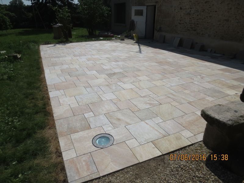 Terrasse en pierre pavée jouxtant une maison, avec un petit luminaire et une pelouse en arrière-plan.