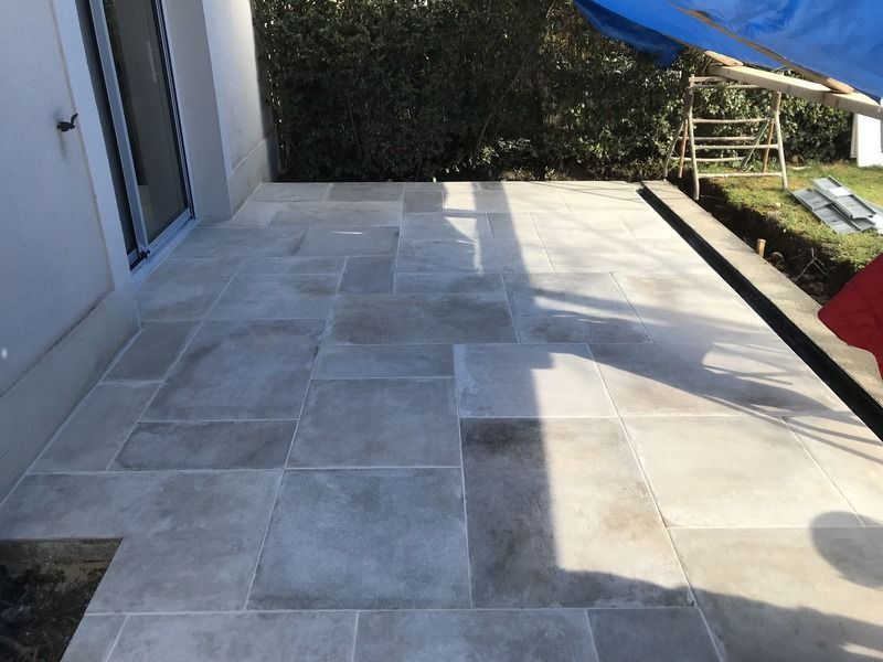 Terrasse carrelée en pierre jouxtant un bâtiment blanc, où le soleil et l'ombre se projettent sur le sol.