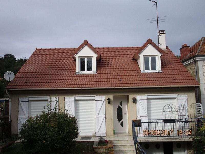 Petite maison blanche au toit de tuiles rouges et aux lucarnes, avec un porche et des volets.
