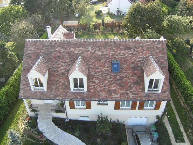 Vue aérienne d'une maison blanche au toit de tuiles rouges, avec des lucarnes et une allée sinueuse traversant des arbustes verts.
