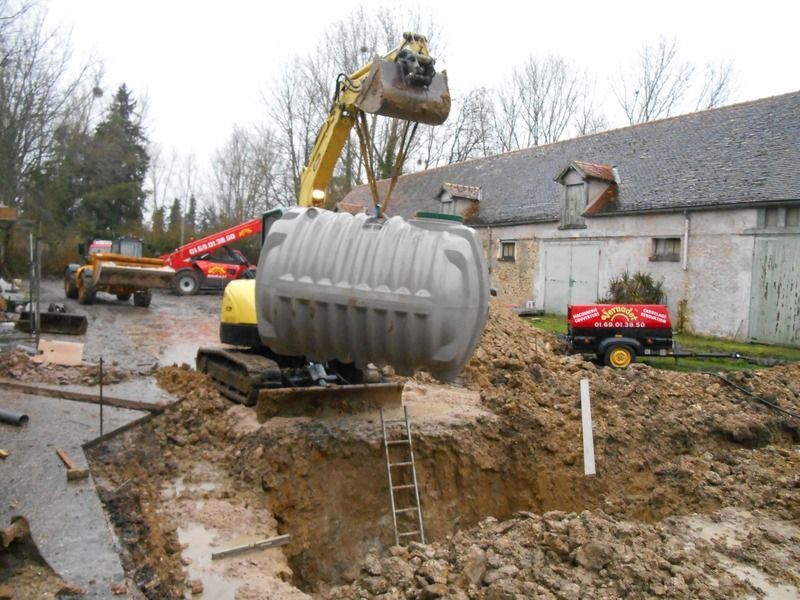 Une excavatrice soulève une grande cuve à eaux grises au-dessus d'un fossé boueux à côté d'un bâtiment rural.