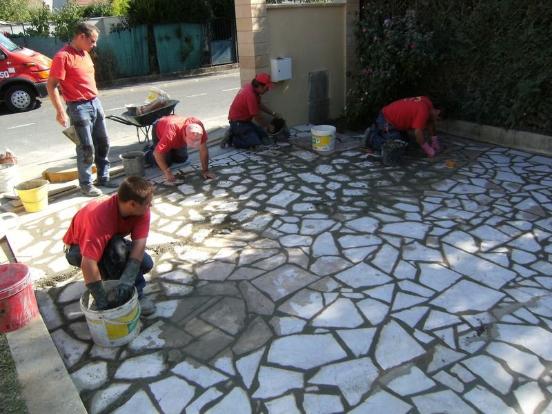 Des ouvriers en chemises rouges posent des pavés sur une terrasse en plein soleil.