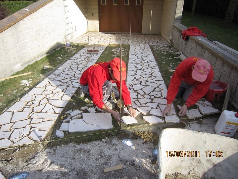 Deux ouvriers sont agenouillés sur une terrasse, posant des pierres grises dans un chemin de béton à côté d'un garage.