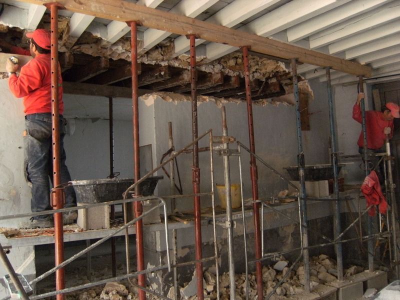 Des ouvriers en vestes rouges démolissent un mur intérieur à l'aide d'échafaudages, des gravats jonchent le sol.