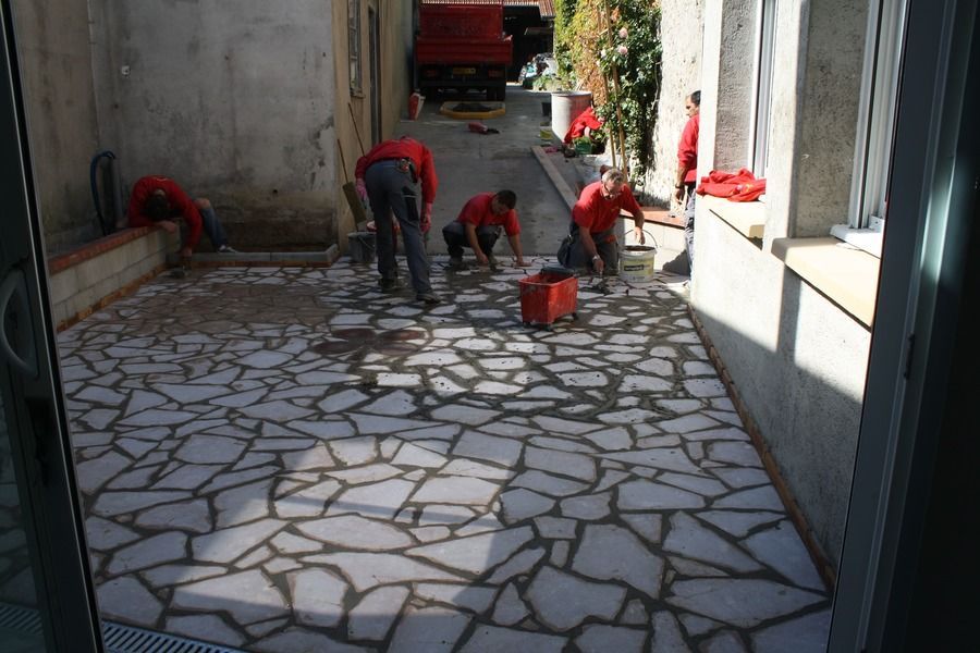 Des ouvriers posent des pavés dans un couloir extérieur, avec des seaux rouges et des outils à proximité.