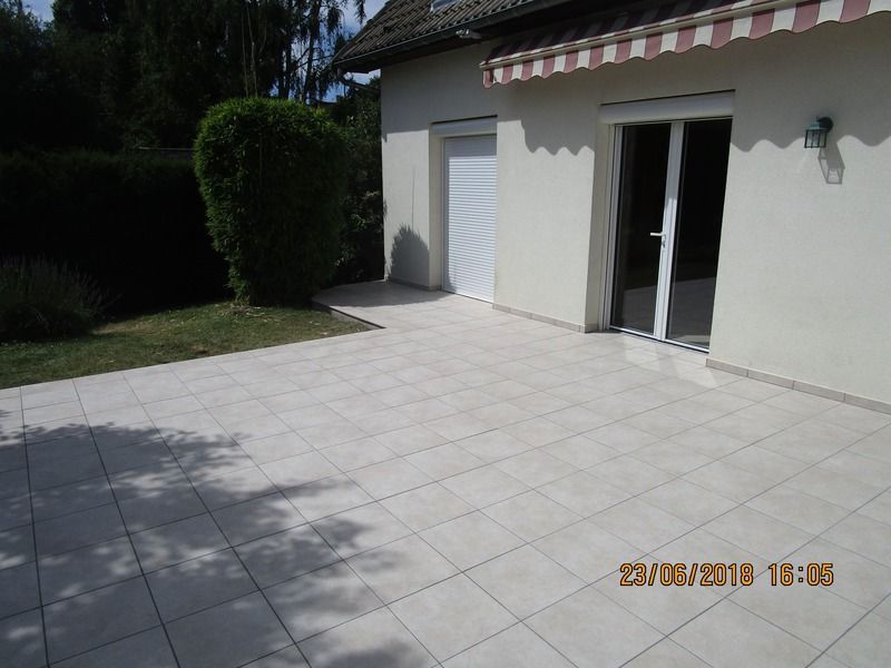 Terrasse en dalles de pierre claire jouxtant une maison blanche, porte vitrée ouverte et store rayé baigné de soleil