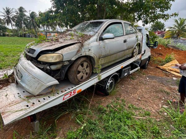 Voiture argentée endommagée, sanglée sur une remorque, dans une zone rurale boueuse.
