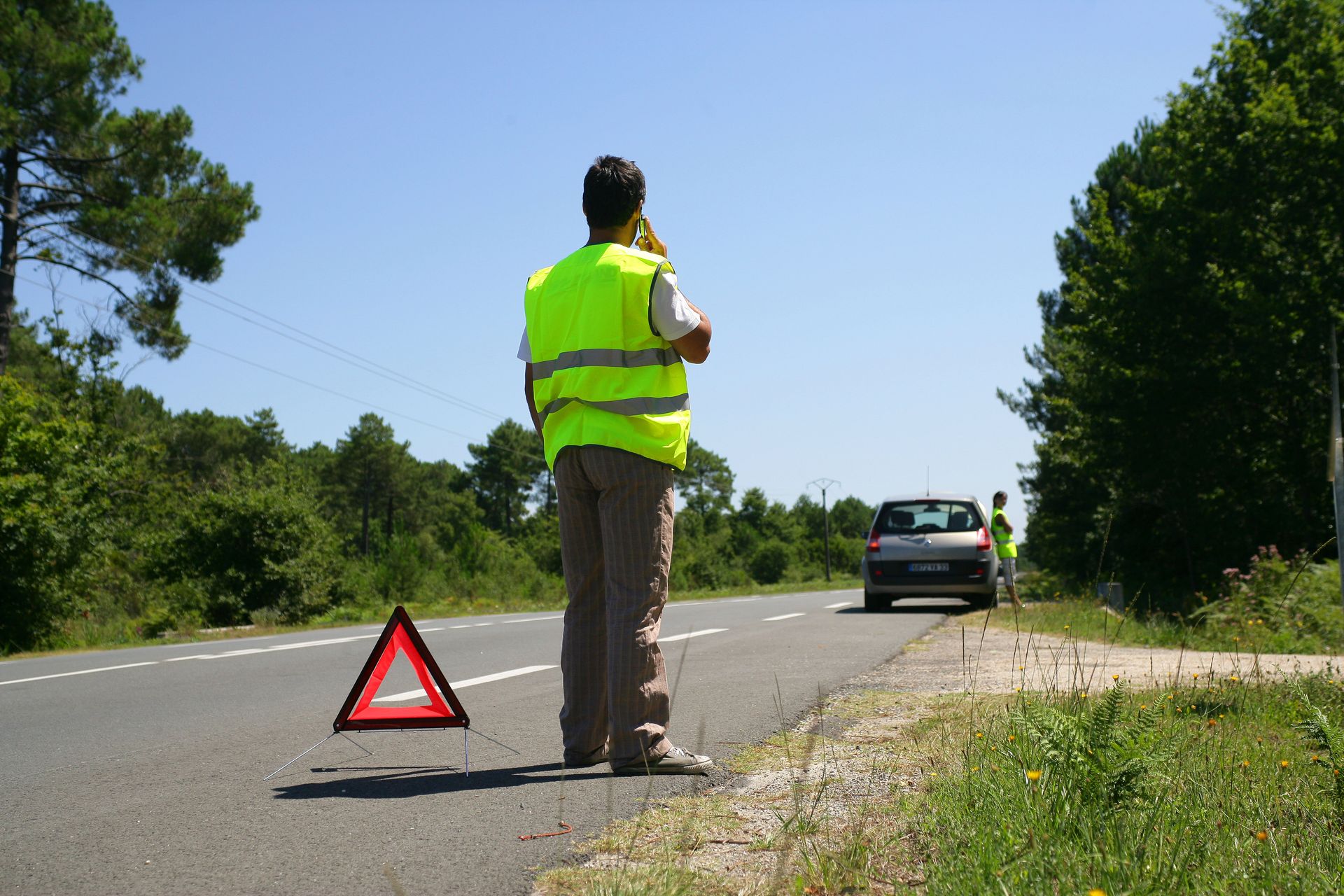 Un homme vêtu d'un gilet fluo, se tient à côté d'un triangle de signalisation, sur une route de campagne près d'une voiture.