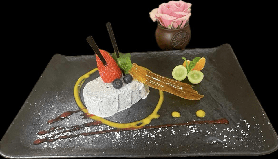 Une assiette noire surmontée d'un dessert et d'un vase de fleurs.