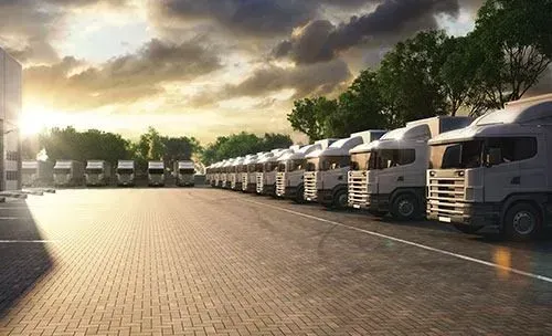 Fila de camiones semirremolques blancos estacionados en un terreno pavimentado al atardecer bajo nubes espectaculares.