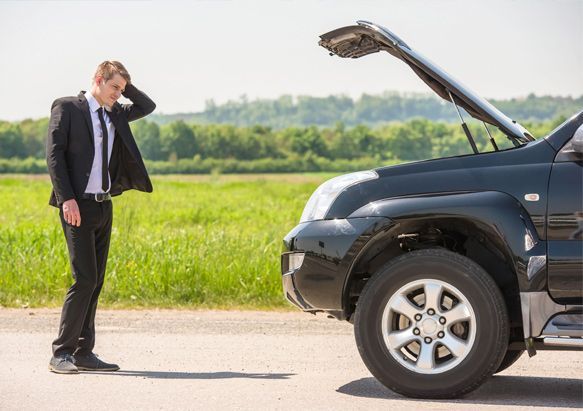 Un hombre con traje y corbata está de pie junto a un coche averiado con el capó abierto.